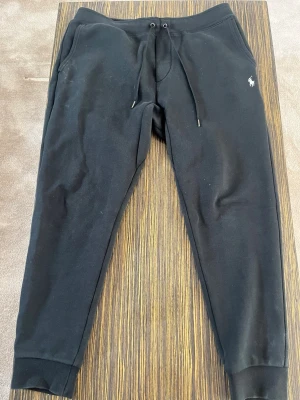 Svarta mjukisbyxor från Polo Ralph Lauren - Snygga svarta mjukisbyxor från Polo Ralph Lauren med dragsko i midjan och diskret logga på vänster höft. Tillverkade i mjukt bomullsmaterial och har muddar vid bensluten. Perfekta för chill dagar eller när du vill vara bekväm.