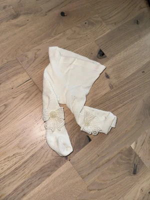 Vita tights med spetsdetalj - Säljer ett par vita tights med dekorativa spetsdetaljer och pärlor vid anklarna. Tightsen har en mjuk och stretchig känsla, perfekta för att ge outfiten en söt touch. Passar dig som gillar romantisk och feminin stil.