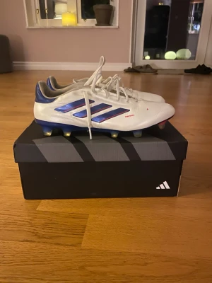 Adidas Copa vita fotbollsskor - Oanvända vita Adidas Copa fotbollsskor med blå detaljer och klassiska tre ränder på sidan. Skorna har snörning, låg profil och fasta dobbar för spel på naturgräs. Tillverkade i syntetmaterial för bra komfort och kontroll på planen.