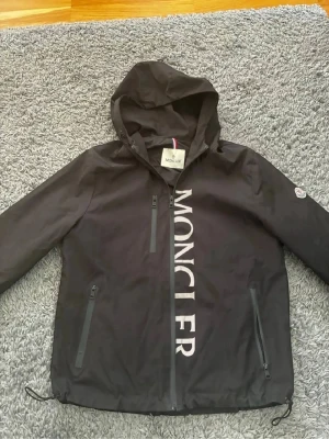 Svart vindjacka från Moncler - Snygg svart vindjacka från Moncler med stor logga längs dragkedjan framtill. Jackan har huva, tre dragkedjeförsedda fickor och justerbar nederkant. Tillverkad i lätt och tåligt material, perfekt för blåsiga dagar. Klassisk Moncler-patch på ärmen.