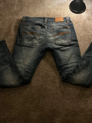 Blå Nudie Jeans grim tim 29-32 - Säljer ett par klassiska blå jeans från Nudie Jeans med snygga slitningar och kontrastsömmar. Jeansen har raka ben, fem fickor och läderpatch bak i midjan. Bakfickorna har Nudie Jeans karaktäristiska broderi. Perfekta för en avslappnad men lite mera grich stil. Dessa jeans är i storleken 29 32 respektive 100 cm i benlängd och 40 cm i mid längd priset är inte hugget i Sten så var vänligen att meddela om frågor. Med vänliga hälsningar Eric