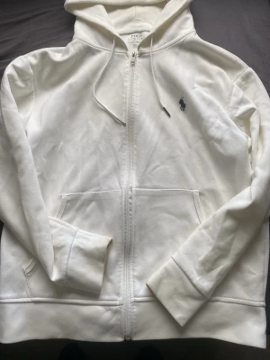 Vit hoodie från Polo Ralph Lauren - Snygg vit hoodie från Polo Ralph Lauren med dragkedja framtill, två fickor och klassisk huva med dragsko. Ikoniska Polo-loggan broderad på bröstet. Tillverkad i mjukt bomullsmaterial som känns riktigt skönt mot huden. Perfekt för en clean och stilren look.