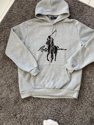 Grå hoodie från Ralph Lauren - Snygg grå hoodie från Ralph Lauren med stor svart polospelare och logga på bröstet. Klassisk känguruficka framtill, ribbade muddar och justerbar dragsko i huvan. Tillverkad i mjukt sweatshirtmaterial som är perfekt för chill dagar.