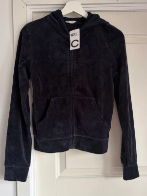 Mörkblå velour hoodie från C - Mjuk och skön mörkblå hoodie i velour från C. Hoodien har dragkedja framtill, känguruficka och huva. Perfekt för en chill och avslappnad stil. Materialet är lent och ger en lyxig känsla. Passar dig som gillar bekväma och snygga plagg.
