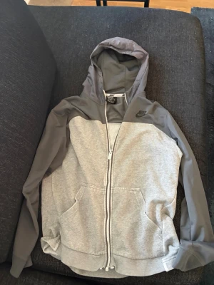 Grå zip hoodie från Nike - Snygg tvåfärgad hoodie från Nike i ljusgrått och mörkgrått med dragkedja framtill. Stor Nike-logga på ryggen och mindre logga på bröstet. Utrustad med huva och två fickor framtill. Perfekt för en sportig och avslappnad stil.
