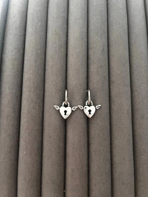 Silverörhängen med hjärtlås och vingar - Säljer ett par små silverfärgade örhängen med hjärtformade hängen som har ett nyckelhål i mitten och små vingar på sidorna. Örhängena har en enkel klickstängning och är tillverkade i metall. Perfekta för dig som gillar unika detaljer och söta smycken.