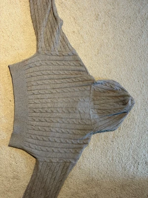 Grå kabelstickad hoodie med dragkedja - Säljer en grå kabelstickad hoodie med dragkedja framtill. Tröjan har ribbade muddar och en mysig huva. Perfekt för dig som gillar en avslappnad men ändå stilren look. Materialet känns mjukt och bekvämt, och den passar lika bra till jeans som till mjukisbyxor.