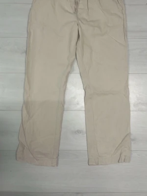 Beige chinos med raka ben - Snygga beige chinos med raka ben och klassisk passform. Byxorna har framfickor och bakfickor, samt bälteshällor i midjan. Tillverkade i ett mjukt bomullstyg som ger en avslappnad look. Perfekta för dig som gillar stilrena och enkla plagg.