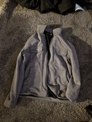 Grå mockajacka med dragkedja - Snygg grå mockajacka med hög krage och dragkedja framtill. Jackan har två stora bröstfickor med lock och en clean, minimalistisk look. Perfekt för dig som gillar stilrena plagg med en modern touch.