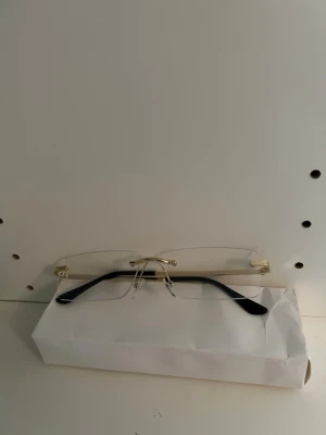 Guldiga glasögon utan bågar - Stilrena glasögon med transparenta, rektangulära linser och tunna guldfärgade skalmar. Skalmarna har svarta ändar och näskuddar i silikon för extra komfort. Perfekt accessoar för en minimalistisk look.