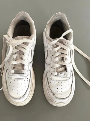 Vita Nike Air Force 1 sneakers - Klassiska vita Nike Air Force 1 sneakers i lågt utförande. Skorna har en chunky sula, perforerad tåbox och ikoniska swoosh-loggan på sidan. Tillverkade i läder med vita skosnören och detaljer. Perfekta för en clean och tidlös streetstil.