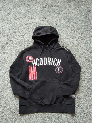 Svart hoodie från Hoodrich med tryck - Svart hoodie från Hoodrich med stor vit logga och röda detaljer på bröstet. Hoodien har huva med dragsko, känguruficka och patchar med text och emblem. Tillverkad i mjuk bomullsblandning och har en avslappnad passform. Perfekt för dig som gillar streetwear.