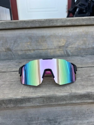 Pastellrosa Sportiga spegelglas solglasögon - Säljer ett par sportiga solglasögon med spegelglas i regnbågsfärger och svart båge. Glasen har en futuristisk, kantig form och ger en cool vibe till din outfit. Perfekta för dig som vill sticka ut och skydda ögonen mot solen.