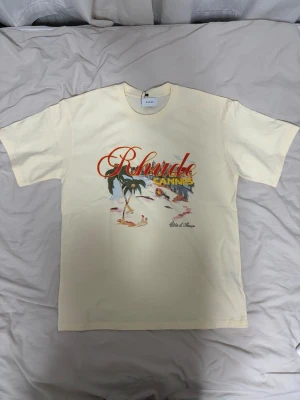 Rhude Cannes t-shirt med tryck - Snygg cremevit t-shirt från Rhude med färgglatt tryck framtill som visar en strandscen, palm och texten 'Cannes' i gult och rött. Rund halsringning och klassisk passform. Perfekt för dig som gillar streetwear och grafiska prints.