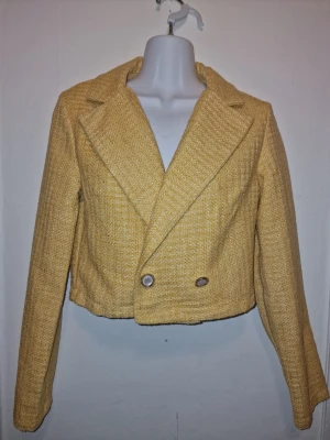 Gul kort kavaj/jacka - Trendig gul kavaj i kort modell med dubbelknäppning och breda slag. Tillverkad i ett grovt, vävt material som ger en cool textur.