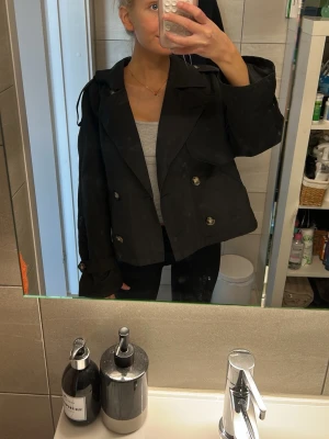Svart trenchcoat från Vero Moda - Kort svart trenchcoat från Vero Moda med dubbelknäppning och stora slag. Jackan har dekorativa axelremmar och knappar vid ärmsluten. Tillverkad i ett slätt tyg som ger en klassisk look. Perfekt för dig som vill ha en stilren och trendig jacka. Köpt för 500kr förra våren, använd fåtal gånger 🩷