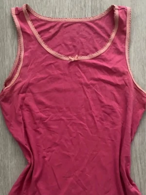 Rosa linnetopp med spetskant - Söt rosa linnetopp med ljusrosa spetskant runt halsringningen och ärmhålen. Liten rosett framtill som detalj. Perfekt att styla lager på lager eller till jeans. Mjuk och skön känsla.