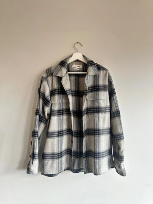 Grå overshirt i ullmix från Samsoe - Overshirt i grått och svart rutigt mönster från Samsoe. Tillverkad i en mjuk ullmix med klassisk krage, knäppning framtill och två bröstfickor. Perfekt lager på lager-plagg för en avslappnad och trendig look.