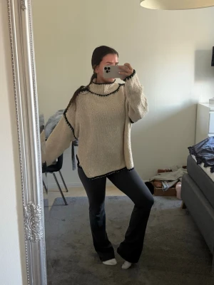 Oversize beige stickad tröja - En oversize stickad tröja i beige med svarta detaljer längs hals, ärmar och nederkant. Tröjan har hög krage och långa ärmar, perfekt för att hålla sig varm. Materialet ser ut att vara mjukt och grovt stickat, vilket ger en avslappnad look.