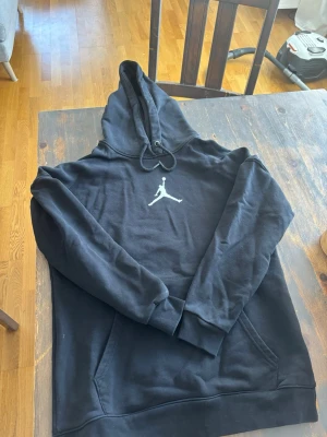 Svart Jordan hoodie med logga - Snygg svart hoodie från Jordan med den klassiska Jumpman-loggan i vitt på bröstet. Hoodien har huva med dragsko och en stor magficka. Tillverkad i mjukt material som känns skönt mot huden. Perfekt för en sportig och avslappnad stil.
