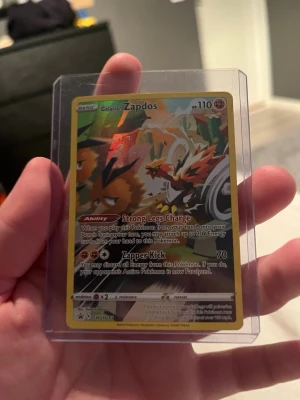 Pokémonkort Galarian Zapdos - Säljer ett Pokémonkort med Galarian Zapdos i färgerna gul, röd, svart och vit. Kortet har holografisk effekt och är inplastat i ett skyddsfodral. Texten på kortet är på engelska och det har 110 HP. Perfekt för samlare eller Pokémonfans.