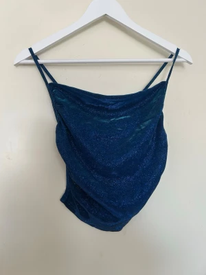 Blå glittrig linnetopp med tunna band - Snygg blå glittrig linnetopp med tunna axelband och draperad framsida. Toppen har en croppad passform och är perfekt för dig som vill sticka ut. Materialet känns mjukt och stretchigt, vilket gör den bekväm att bära.