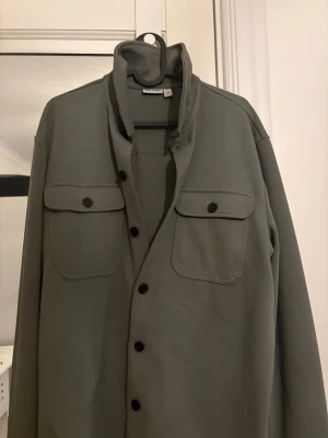 Grön overshirt från Only & Sons - Stilren grön overshirt från Only & Sons med klassisk krage, svarta knappar och två bröstfickor med lock. Tillverkad i ett strukturerat tyg som ger en modern look. Perfekt att bära öppen över en t-shirt eller stängd för en mer dressad stil. Använts få gånger den är som ny 