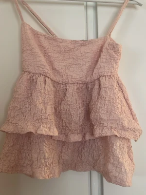 Ljusrosa volangig linnetopp - Söt ljusrosa linnetopp med tunna axelband och dubbla volanger. Toppen har en skrynklig struktur som ger en avslappnad och trendig look. Perfekt för varma dagar och enkel att styla med jeans eller kjol.