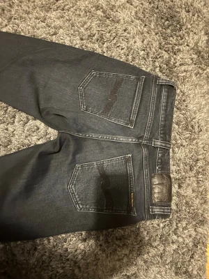 Mörkblå jeans från Nudie Jeans - Snygga mörkblå jeans från Nudie Jeans med klassisk femficksdesign och kontrastsömmar. Bakfickorna har Nudie Jeans karakteristiska broderi och ett brunt läderpatch i midjan. Tillverkade i slitstark denim, perfekta för en avslappnad och trendig look.