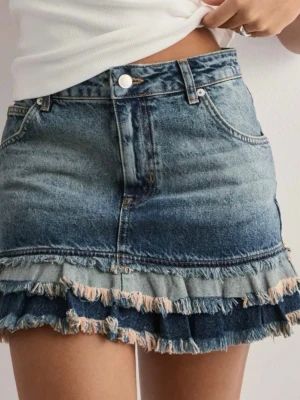 Blå jeanskjol med volang och fransar - Trendig blå jeanskjol med volangkant och slitna fransar längst ner. Kjolen har klassisk femficksdesign, knapp och dragkedja framtill samt en snygg tvättad denimlook. Perfekt för dig som gillar en cool och avslappnad stil.