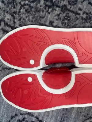 Louboutin skor - Stilrena svarta sneakers med vit och röd sula. Ovandelen är i svart konstläder med matchande svarta skosnören. Sulan har en unik röd undersida med grafiskt mönster och vit kant. Perfekta för dig som vill ha en clean look med en twist.