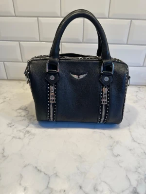 Svart handväska med nitar och detaljer Zadig & Voltaire - Snygg svart handväska i skinnimitation med silverfärgade metalldetaljer, dubbla handtag och dekorativa nitar längs kanterna. Väskan har två dragkedjor framtill och ett emblem i metall. Perfekt för dig som gillar en edgy och trendig look.