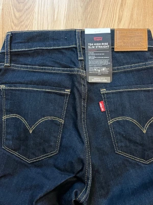 Levi's High Rise Straight Jeans blå - Mörkblå raka jeans från Levi's med hög midja och klassiska kontrastsömmar. Femficksmodell med knappgylf och detaljer i kopparfärg. Tillverkade i slitstark denim, perfekta för en clean och tidlös look. Storlek W25 L32