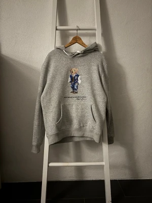 Grå Polo Ralph Lauren hoodie - Säljer en grå hoodie från Polo Ralph Lauren med den ikoniska björnen på bröstet. Hoodien har huva med vita snören. Tillverkad i mjukt material som är riktigt skönt. Perfekt för dig som gillar sköna kläder. Storleken är (M) men är lite mindre i storleken. Skriv gärna om du undrar något.