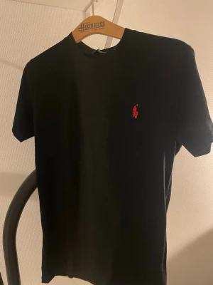 Svart t-shirt från Ralph Lauren - Klassisk svart t-shirt från Ralph Lauren med röd broderad logga på bröstet. T-shirten har rund halsringning och korta ärmar. Perfekt basic-plagg med stilren design. (passar mer Som Medium)