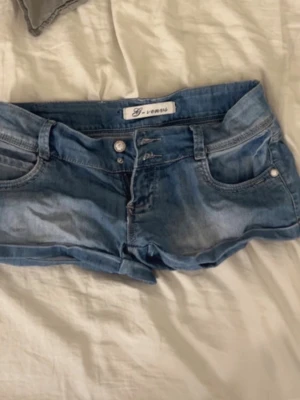 Blå denimshorts från G-venus - Säljer ett par ljusblå denimshorts från G-venus med klassisk femficksdesign, knappgylf och bälteshällor. Shortsen har bakfickor med dekorativa sömmar och små märkespatchar. Perfekta för varma dagar och enkel att matcha med allt. OBS: knappen nere är av