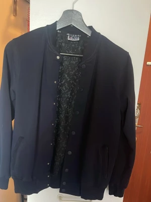 Svart bomberjacka från Cessia - Stilren svart bomberjacka från Cessia med klassisk rund halsringning och tryckknappar framtill. Jackan har ribbade muddar vid ärmslut och nederkant samt två fickor med dragkedja. Insidan har ett mönster med vita bokstäver. Perfekt för en cool och avslappnad look.