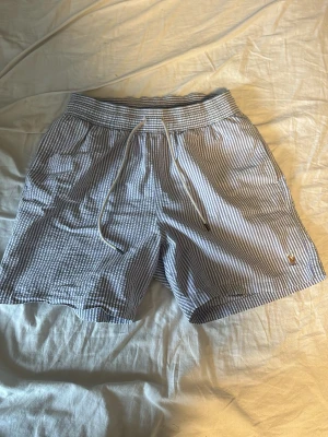 Ralph lauren badbyxor - Säljer ett par snygga randiga badbyxor från ralph lauren i blått och vitt. De har elastisk midja med snörning, sidofickor och en bakficka med knapp. Perfekta för sommaren och tillverkade i mjuk bomull för maximal komfort. Kom med frågor och prisförslag!