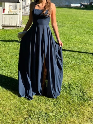 Marinblå långklänning med slits - Säljer en elegant marinblå långklänning med slits och smala axelband. Klänningen har en svepande kjol och en enkel, stilren överdel med lätt drapering. Perfekt för studentbal eller niornas bal. Materialet ger ett snyggt fall. 