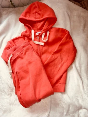 Korallfärgat mjukisset med huva - Säljer ett mjukis- set i korallfärg med matchande hoodie och byxor. Hoodien har dragkedja, stora fickor och vita snören i huvan. Byxorna har resår i midjan och vita dragsnören. Perfekt för chill dagar hemma. Inte använd bara provad. Kontakta gärna innan köp eller vid funderingar. Kan diskutera priset.