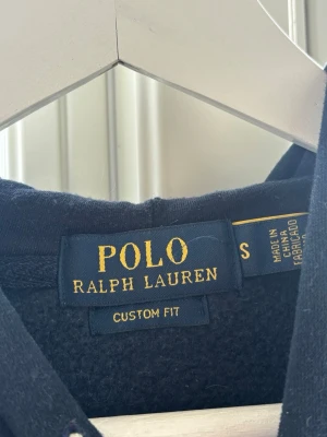 Ralph Lauren hoodie  - Mörkblå hoodie från Polo Ralph Lauren med vita snören och röd broderad logga på bröstet. Klassisk custom fit-modell med huva och mjukt material på insidan. Perfekt för en avslappnad och stilren look.