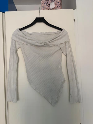 Vit asymmetrisk topp med pärldetalj - Snygg vit topp med asymmetrisk nederkant och offshoulder-design. Toppen har ribbat tyg och en pärldetalj framtill vid halsringningen. Långärmad och perfekt för dig som vill sticka ut med en unik stil.