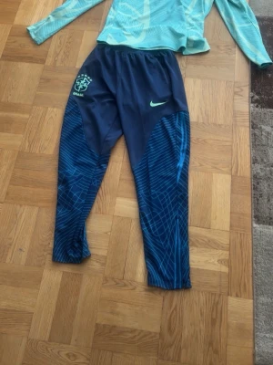 Nike Brasil träningsset blå/turkos - Snyggt träningsset från Nike med Brasil-logga. Tröjan är turkos med grafiskt mönster och halv dragkedja, långärmad. Byxorna är mörkblå med ljusblå detaljer och elastisk midja. Perfekt för fotbollsträning eller sportaktiviteter.