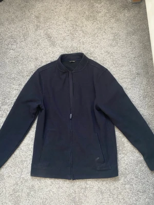 Mörkblå ziptröja från Massimo Dutti - Mörkblå ziptröja från Massimo Dutti med hög krage och långa ärmar. Tröjan har två fickor framtill och är tillverkad i ett mjukt material som passar perfekt för lager-på-lager. Enkel och stilren design.