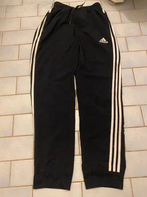 Svarta Adidas mjukisbyxor med ränder - Klassiska svarta mjukisbyxor från Adidas med vita ränder längs sidorna och broderad logga på låret. Byxorna har resår och snörning i midjan samt muddar vid bensluten. Perfekta för chill eller träning.