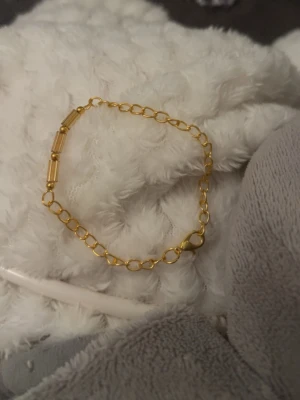 Tunt guldarmband  -  armband i guldfärgad metall med små stavformade länkar och tunn kedja. 