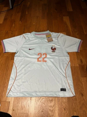 France wc 26 away kit Ekitike #22 - Snygg vit fotbollströja från Nike med Frankrikes landslagsmärke och nummer 22 i orange på bröstet och ryggen. Tröjan har röda, blå och vita detaljer på ärmsluten samt namnet Ekitike på ryggen. Perfekt för fotbollsfans som vill sticka ut på planen.