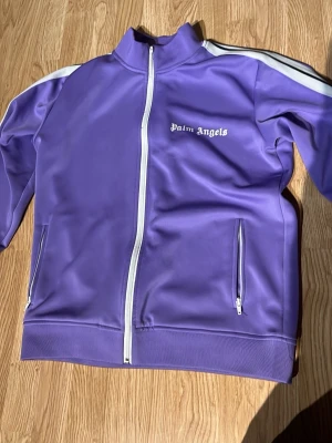 Lila track jacket från Palm Angels - Säljer en lila track jacket från Palm Angels med vit dragkedja och vita ränder längs ärmarna. Jackan har hög krage, två fickor med dragkedja och logga på bröstet. Perfekt för en sportig och trendig look.