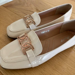 Beige loafers med gulddetalj - Snygga beige loafers i lackat material och dekorativ guldfärgad kedjedetalj framtill. Skorna har låg sula och stilren design som passar till många olika outfits. Storlek 38! Så trendiga till sommaren 