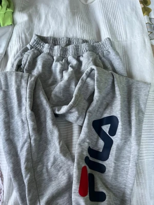 Gråa mjukisbyxor från Fila - Säljer ett par gråa mjukisbyxor från Fila med stor logga i marinblått och rött på benet. Byxorna har resår i midjan och sidofickor. Perfekta för chill dagar eller träning. Mjukt och bekvämt material. Barn storlek  11-12 år